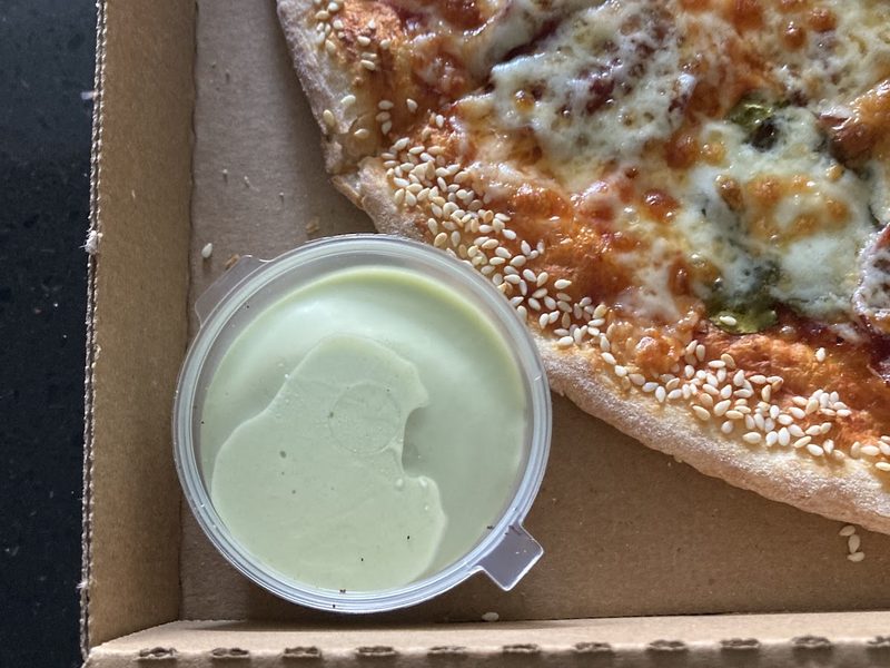 A pizza in an open cardboard box with a large tub of dip in the foreground