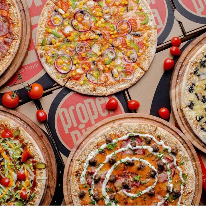 An overhead spread of several whole pizzas on red serving boards with tomatoes and peppers